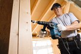 Person wearing safety equipment drills into a wooden beam with a rotary hammer.