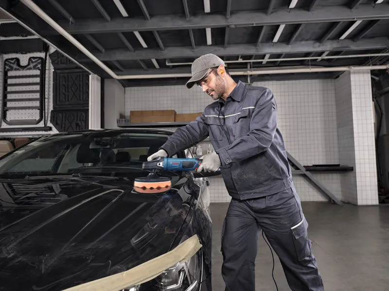 A person wearing safety equipment polishes a car hood with a power tool.