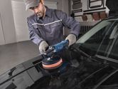 A person wearing safety equipment polishes a car hood with a power polisher.