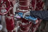 A person wearing safety equipment uses a cordless ratchet wrench on industrial piping.