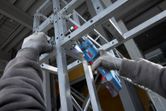 A person wearing safety equipment tightens bolts on a metal frame using a cordless ratchet wrench.