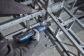 Person wearing safety equipment tightens bolts on a metal frame using a cordless ratchet wrench.