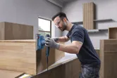 A person wearing safety equipment sands a wooden cabinet with an orbital sander.