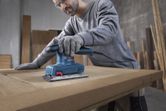 A person wearing safety equipment sands a wooden door with an orbital sander.