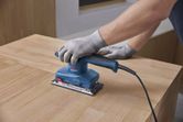 A person wearing safety equipment uses an orbital sander on a wooden surface.