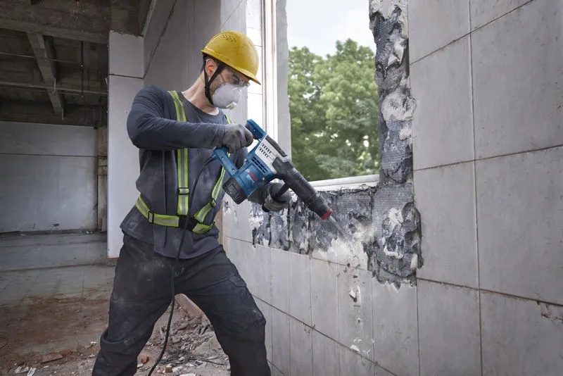 A worker wearing safety equipment uses a demolition hammer to break a tiled wall.