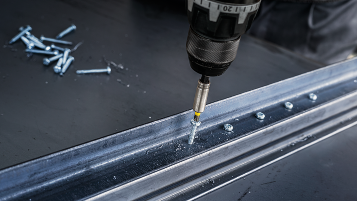 Cordless drill driving a screw into a metal beam on a workshop table.