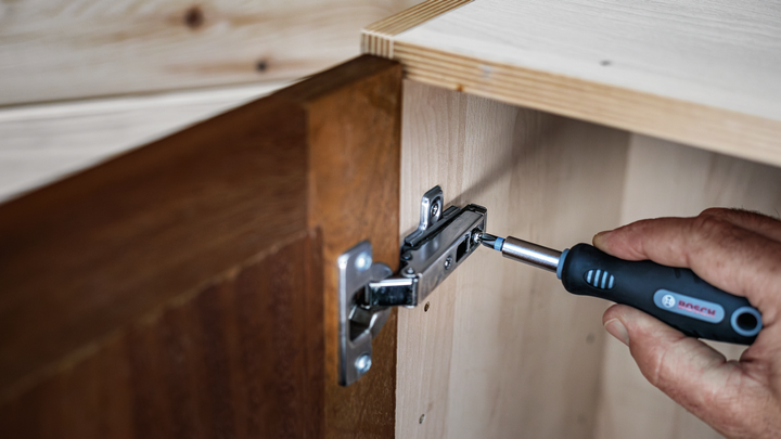 Person using a screwdriver to tighten a hinge on a wooden cabinet door.