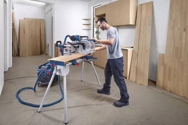 A person wearing safety equipment cuts a wooden board using a mitre saw in a workshop.