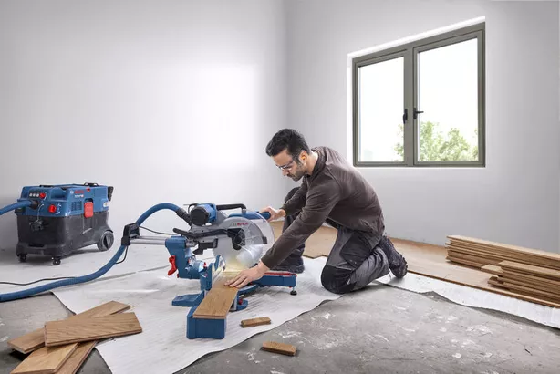 A person wearing safety equipment cuts wood planks with a mitre saw in a bright room.