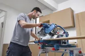 A person wearing safety equipment cuts wood with a mitre saw in a workshop.