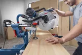 A person operates a mitre saw to cut a wooden board in a workshop.