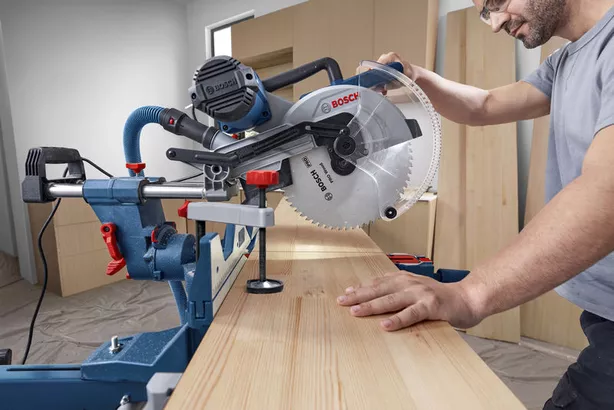 A person operates a mitre saw to cut a wooden board in a workshop.