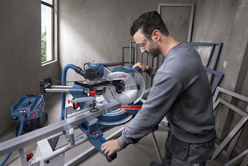 A person wearing safety equipment operates a mitre saw to cut a metal piece.
