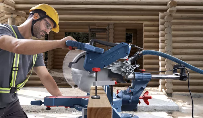A person wearing safety equipment operates a mitre saw to cut wood at a construction site.