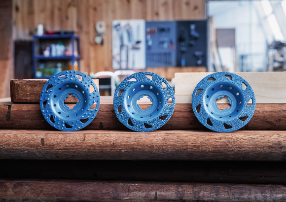 Object #200557594: 7425288_APP_WIDE_2 Three blue grinding wheels are displayed on stacked wooden beams in a workshop.
