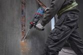 A person wearing safety equipment uses a rotary hammer to chisel concrete from a wall.