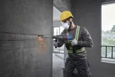 A person wearing safety equipment drills into a concrete wall with a rotary hammer.