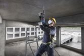 A person wearing safety equipment drills into a concrete ceiling with a rotary hammer.