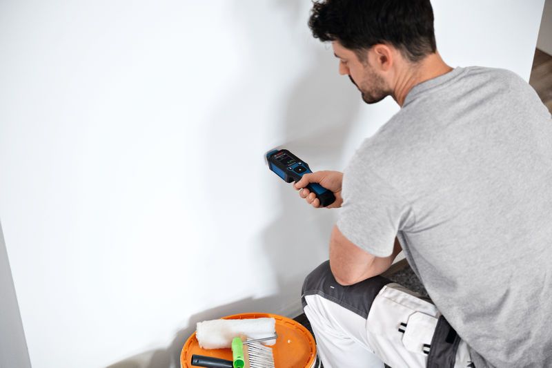 A person uses a material moisture meter against a white wall.