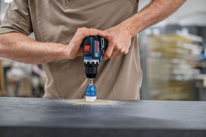 Person operating a cordless drill with a hole saw attachment on a dark surface.