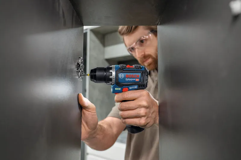 Person wearing safety equipment uses a cordless drill to install a cabinet hinge.