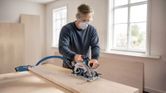 Person wearing safety equipment cuts wood with a cordless circular saw on a workbench.