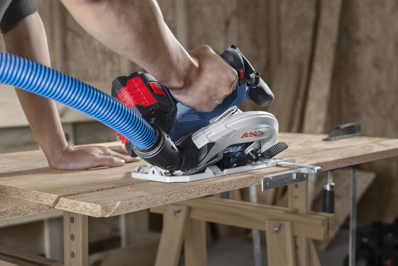 A person guides a cordless circular saw with dust extraction hose while cutting plywood.