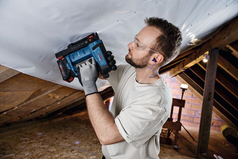A person wearing safety equipment fastens material to a ceiling using a cordless tool.