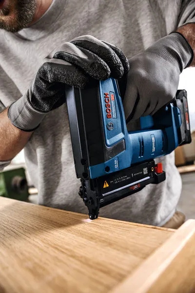 A person wearing safety equipment uses a cordless nail gun on a wooden surface.