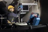 A person wearing safety equipment uses a cordless power tool under a bright jobsite light.