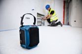 Bluetooth speaker on a construction site floor, person in background wearing safety equipment.