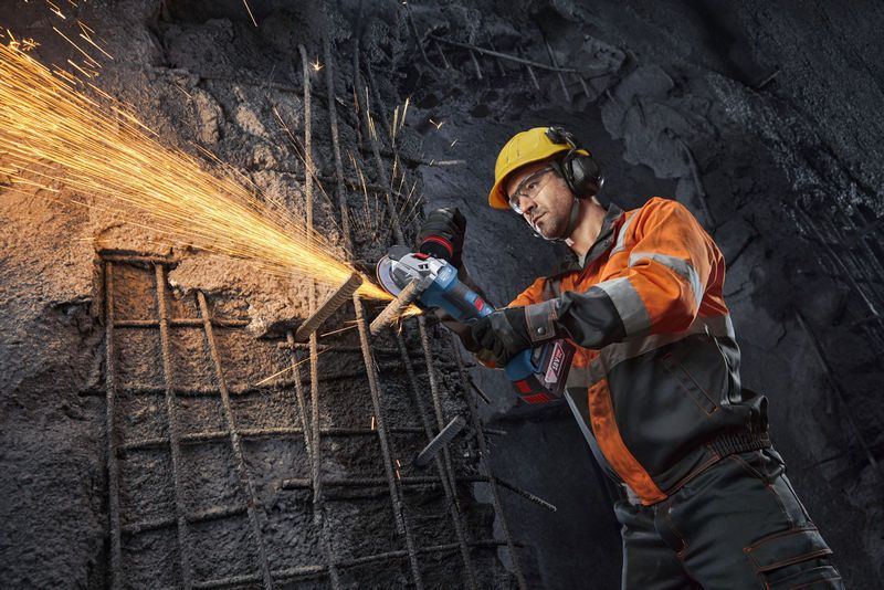 A person wearing safety equipment uses an angle grinder to cut rebar, creating sparks.