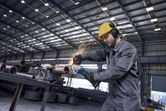 A person wearing safety equipment uses an angle grinder on a metal pipe, creating sparks.
