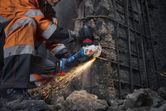 A person wearing safety equipment cuts concrete with an angle grinder, producing sparks.