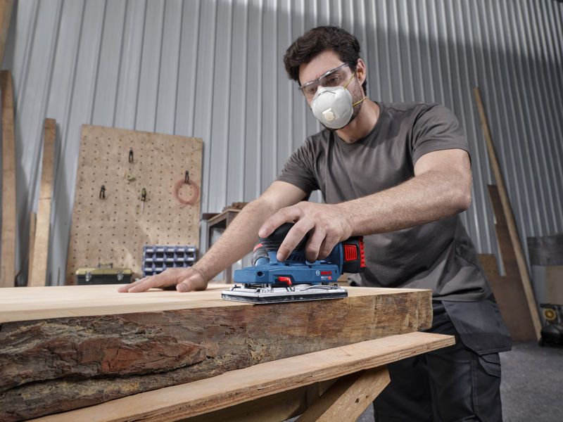 A person wearing safety equipment sands a wooden board with a cordless orbital sander.