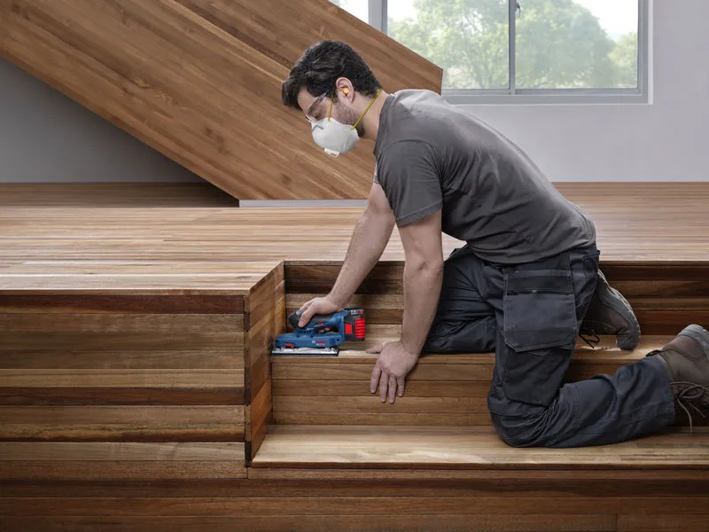A person wearing safety equipment sands wooden stairs with an orbital sander.