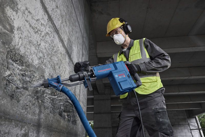 A person wearing safety equipment uses a demolition hammer to break concrete.