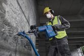 A person wearing safety equipment uses a demolition hammer to break concrete.