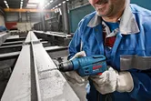 A person wearing safety equipment drills into a steel beam using a blue power tool.
