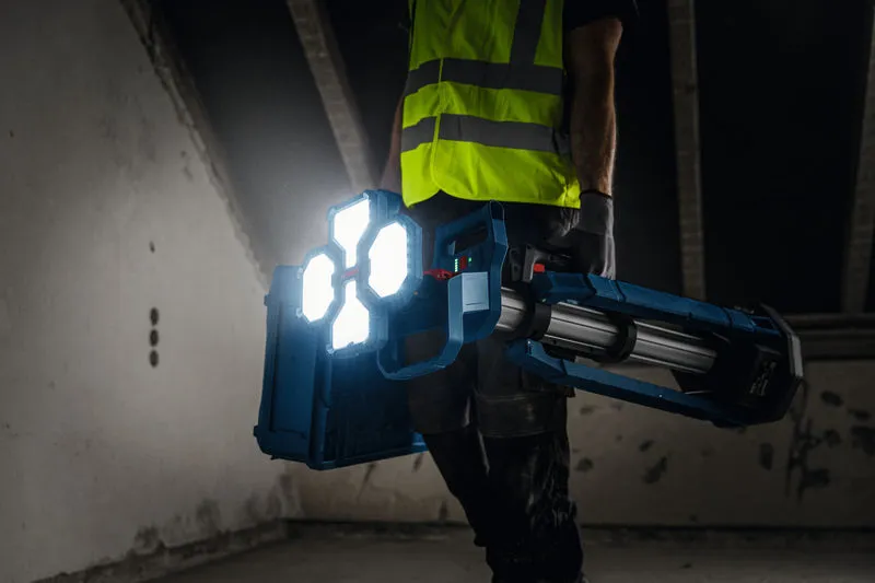 A person wearing safety equipment carries a cordless jobsite light in a dimly lit room.