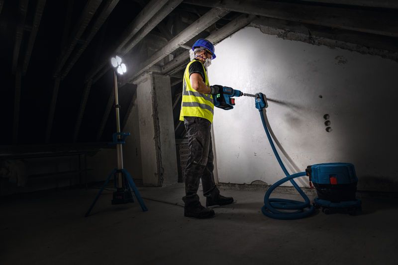 A person wearing safety equipment drills into a wall under a cordless jobsite light.