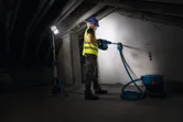 A person wearing safety equipment drills into a wall under a cordless jobsite light.