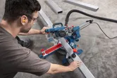 Person wearing safety equipment cuts metal tubing with a mitre saw on a workshop floor.