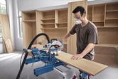 A person wearing safety equipment uses a mitre saw to cut a wooden board.