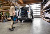 A person wearing safety equipment carries a cordless dust extractor toward an open work van.
