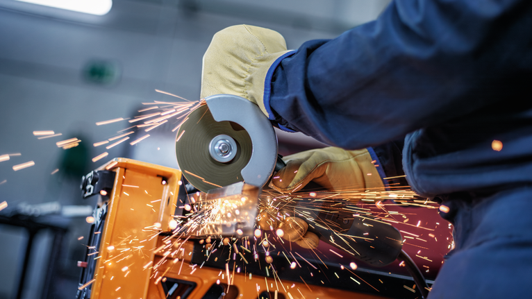 Person wearing safety equipment uses an angle grinder, producing bright sparks.