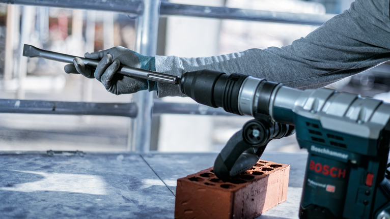 Person wearing safety equipment uses a rotary hammer drill to chisel a red brick.
