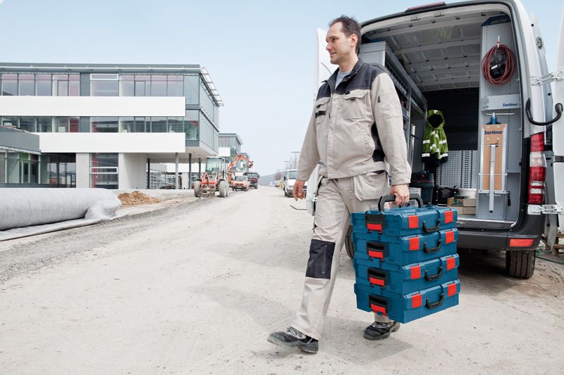A person wearing safety equipment carries stacked toolboxes near a work van at a construction site.