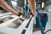 A person operates a power router on a wooden panel connected to a wet and dry extractor.